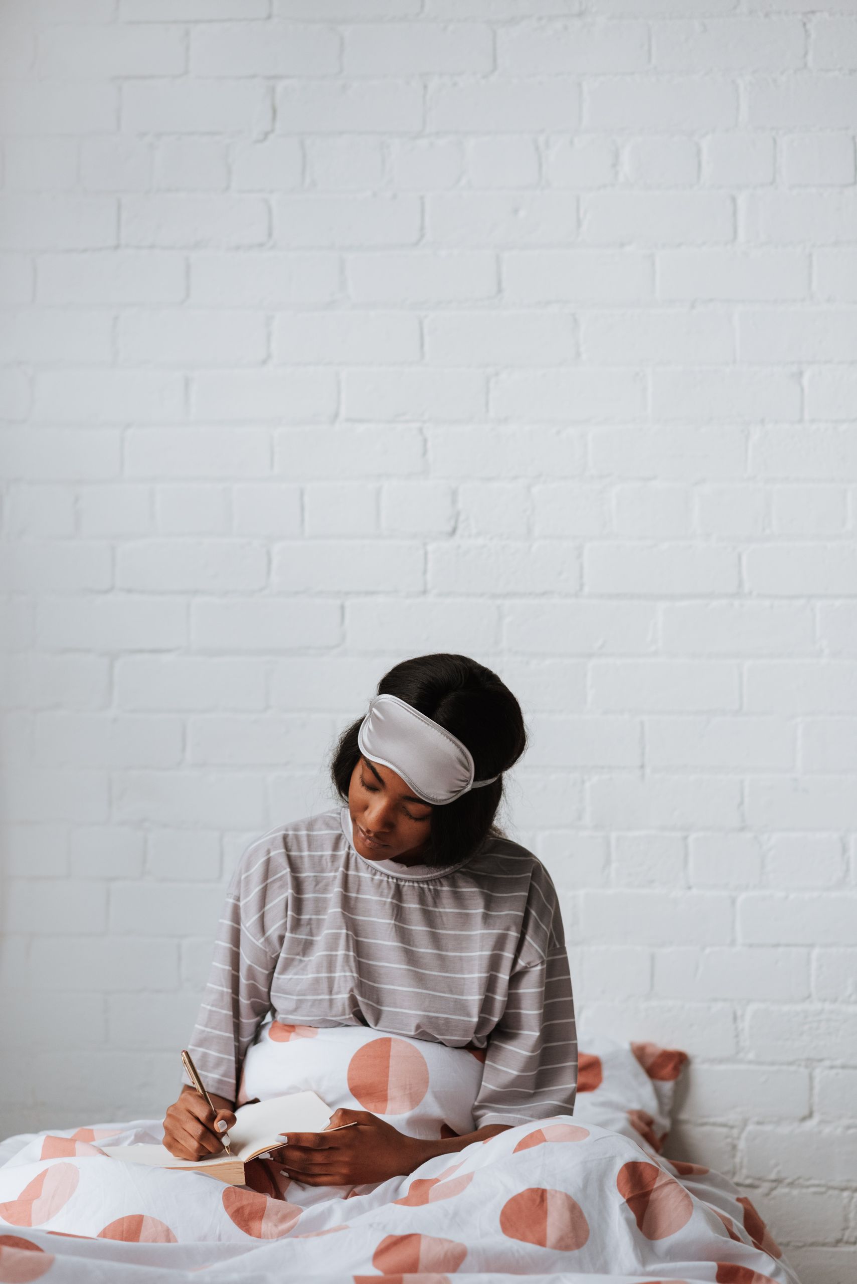 A young woman in striped pajamas and a sleep mask writes in a notebook while sitting on a bed with a polka-dot cover.