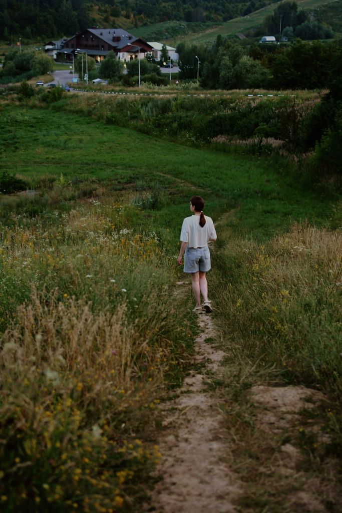 A young woman walks alone on a dirt path through verdant countryside.