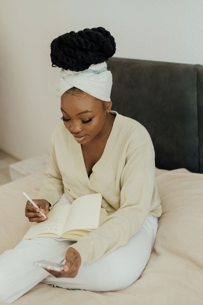 African woman in headwrap sitting on bed, taking notes and using smartphone, in a lifestyle setting.