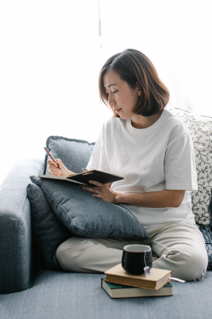 Asian woman in white t-shirt journaling on blue couch, enjoying a peaceful moment indoors.