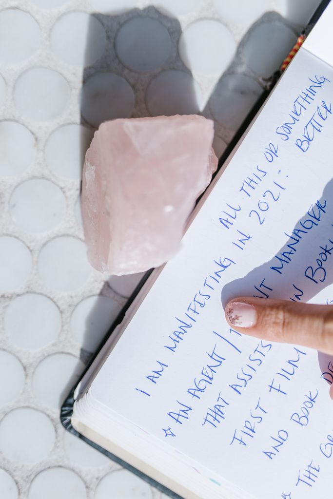 Close-up of manifestation journal prompts page with a rose quartz crystal and finger pointing. 