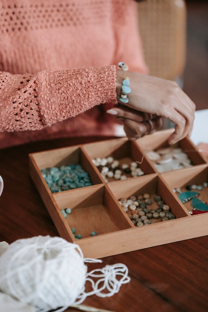 Crop faceless female wearing orange pullover trying on handmade bracelet of blue natural stones and beads sitting at table with box for needlework