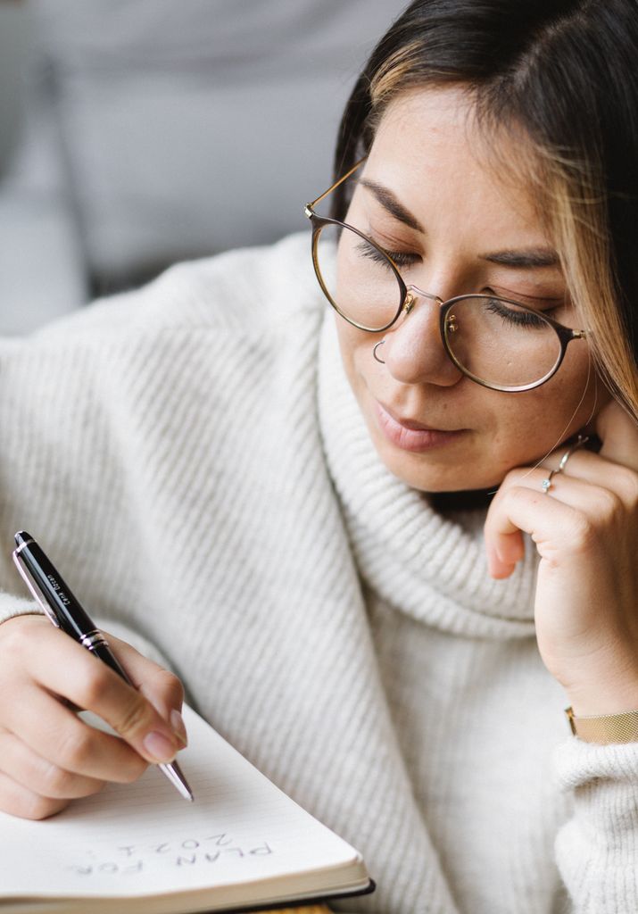 Crop smart female young student in warm casual sweater and eyeglasses writing in notebook while doing year end reflection