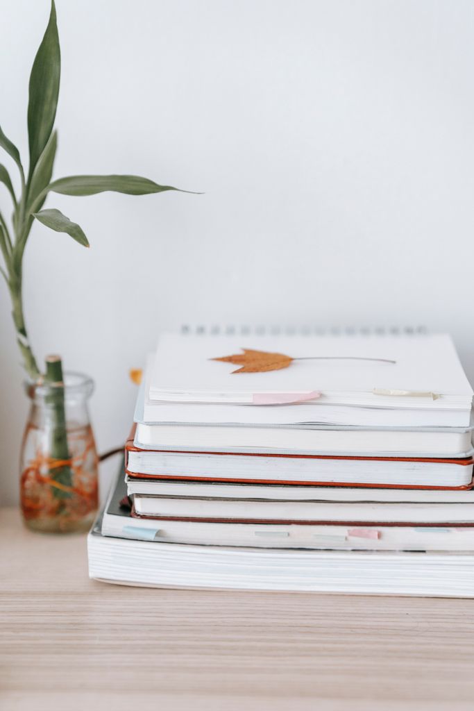 Faded leaf on heap of notebooks against glass vase with lucky bamboo in water on desk on white background