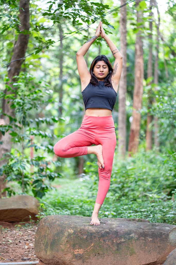 Female doing yoga tree pose on a rock in a lush forest setting, promoting mindfulness and fitness.