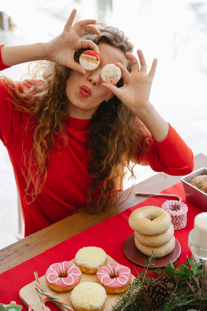 Female with pouting lips covering eyes with tasty gingerbread biscuits at table with assorted doughnuts and bagels during New Year holiday