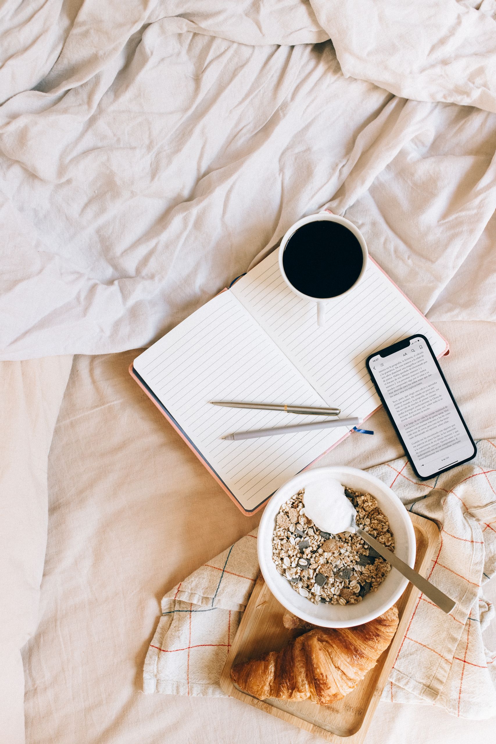 Flat lay of breakfast with coffee, granola, and croissant on a bed for a morning gratitude journal
