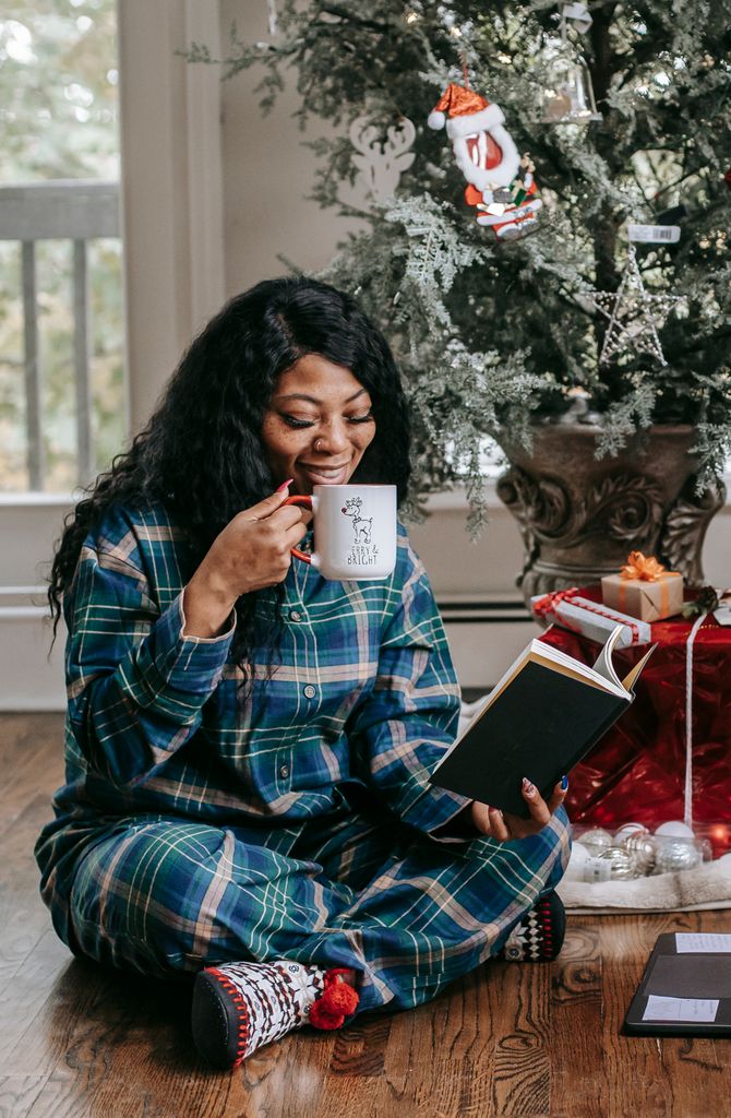 Full body of positive African American female in pajama reading book while sitting on floor under Christmas tree with mug of hot drink