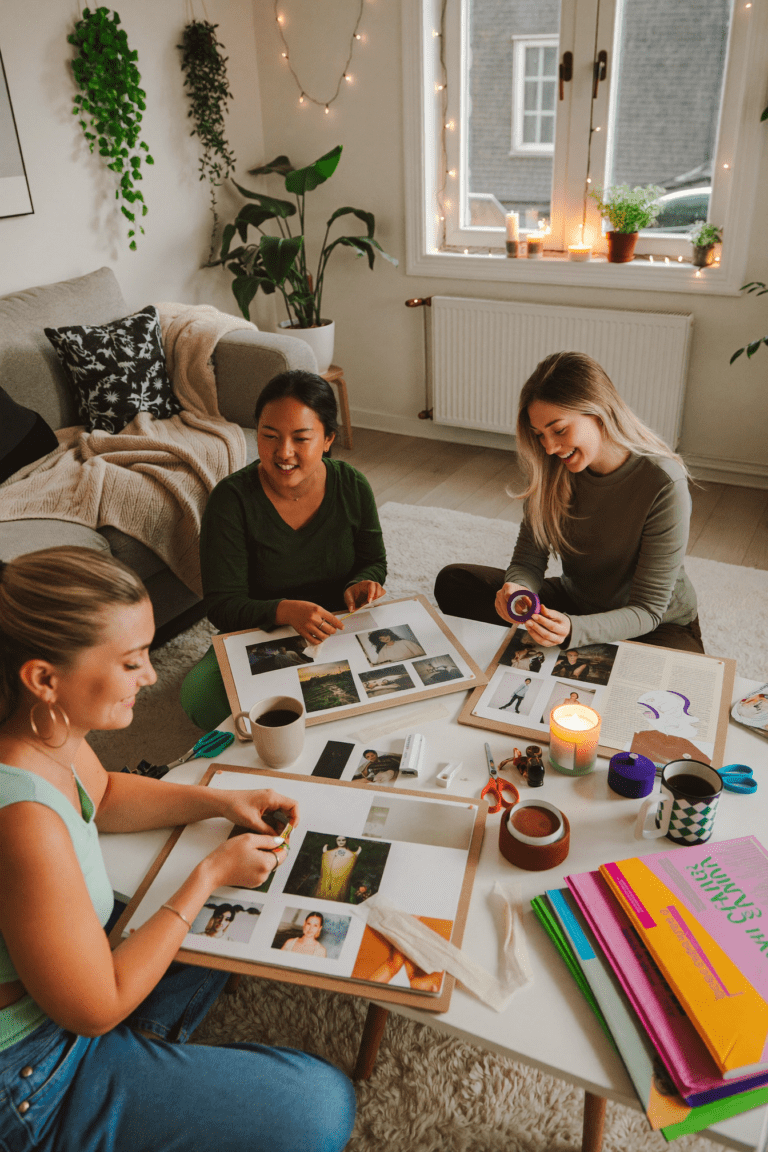 Three women sitting together in a cozy living room at a vision board making party, cutting out magazine clippings and gluing them onto poster boards surrounded by candles, plants, and coffee.
