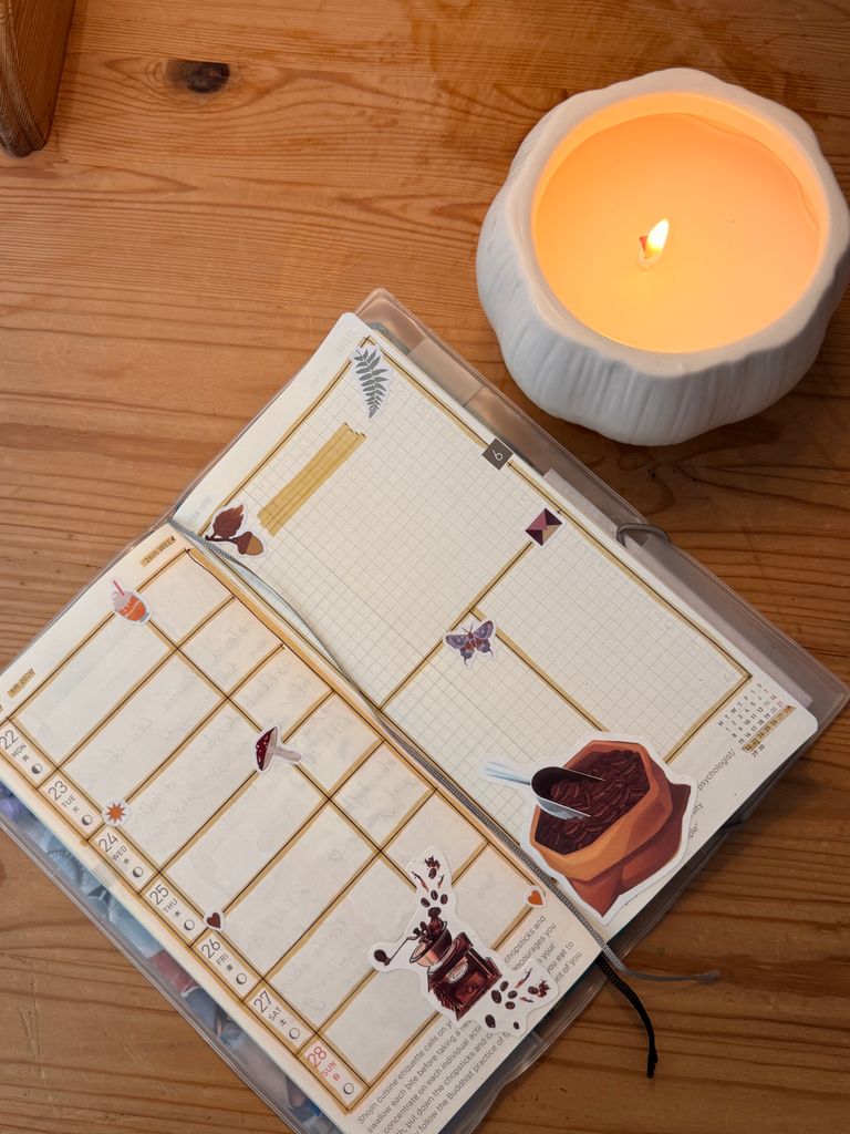 Open journal with coffee-themed stickers beside a lit candle on a wooden table.