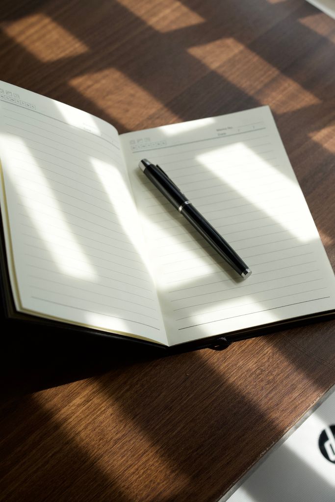 Open notebook with pen on a sunny wooden desk, casting shadows, creating a peaceful workspace vibe.