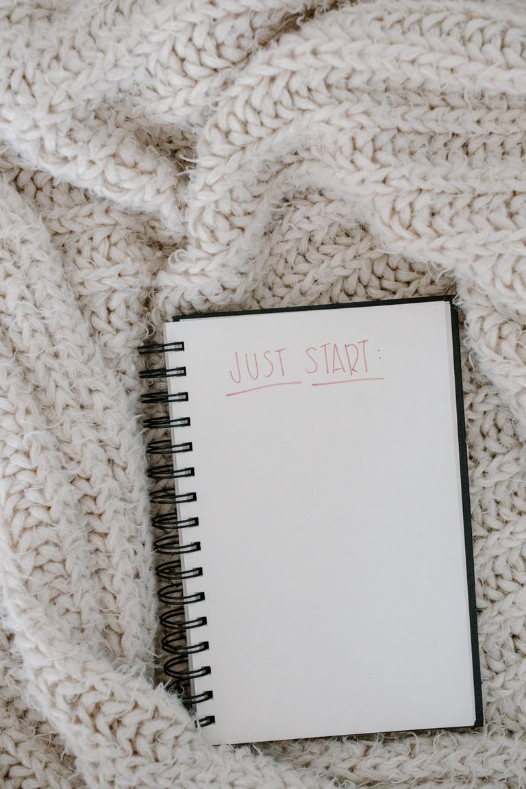 Open spiral notebook on a cozy knitted blanket with 'Just Start' written.
