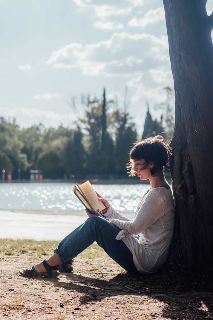 Peaceful scene of a woman reading under a tree by a lake in a sunny park. best books for self-love