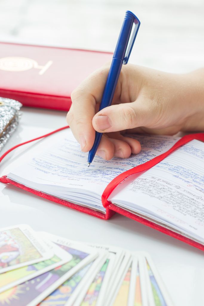 Person writing in a red notebook with tarot cards and pen in view.