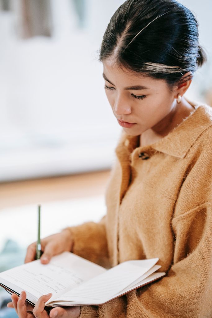 Side view of young Hispanic female in casual clothes with pen reading information from notebook in light room