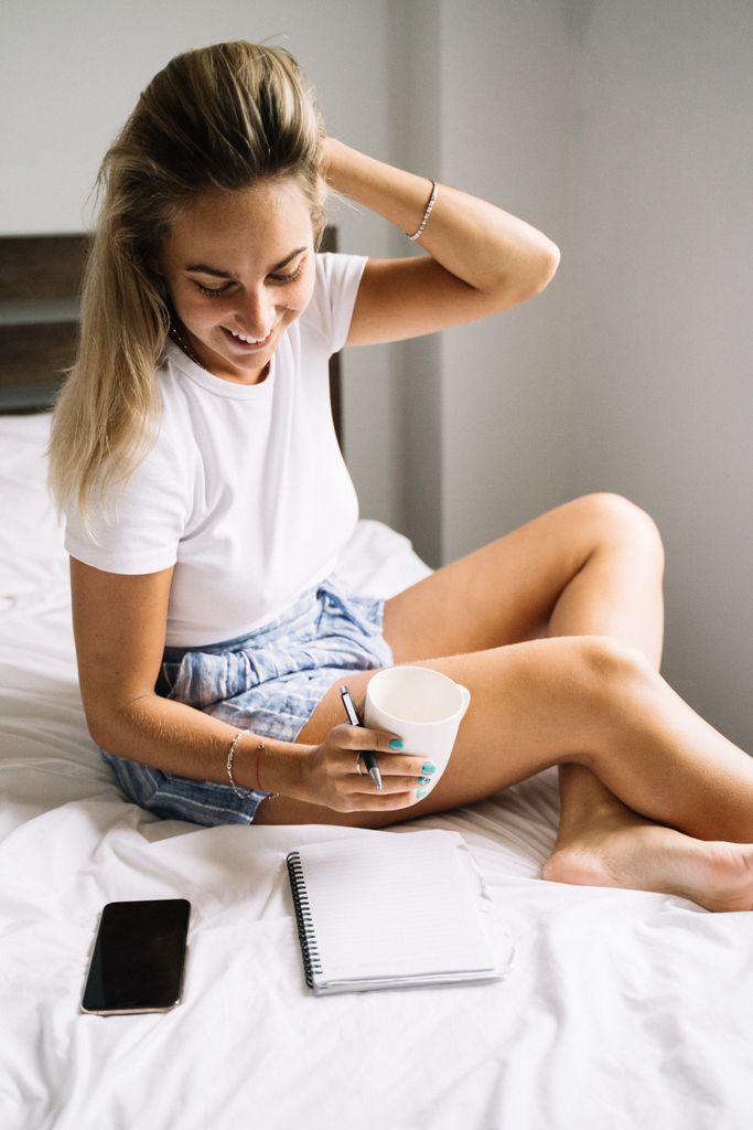 Smiling woman holding a mug while sitting on bed with notebook and phone.