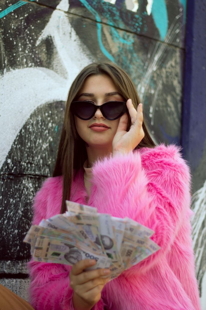 Stylish woman in sunglasses wearing a pink fur coat, holding a stack of cash on an urban street.