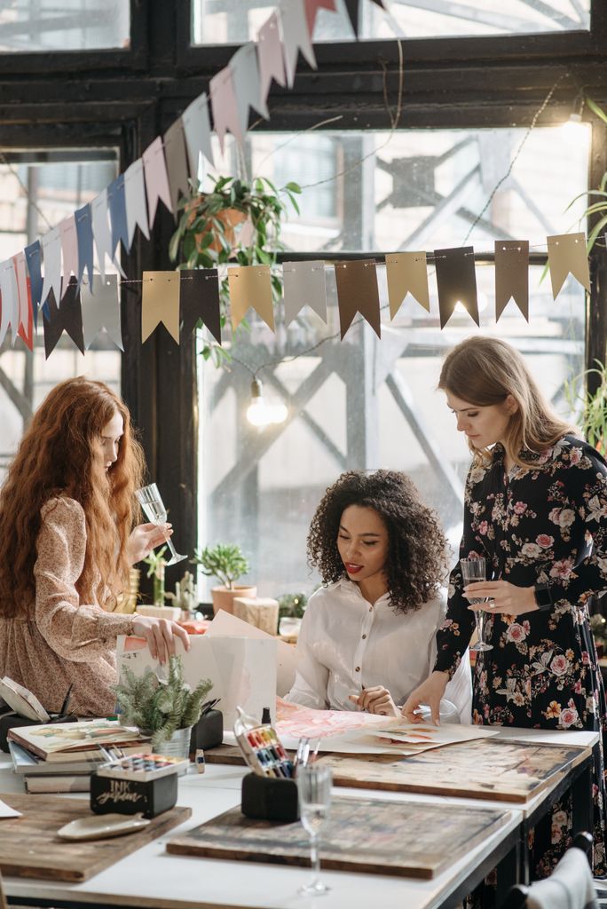 Three women engage in a creative workshop indoors, surrounded by art supplies and festive decor.