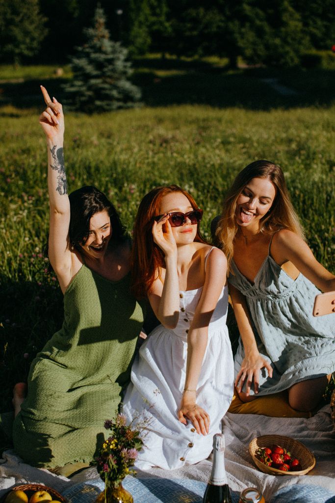 Three women enjoying a sunny picnic with drinks and fruits. Perfect for lifestyle and fashion.