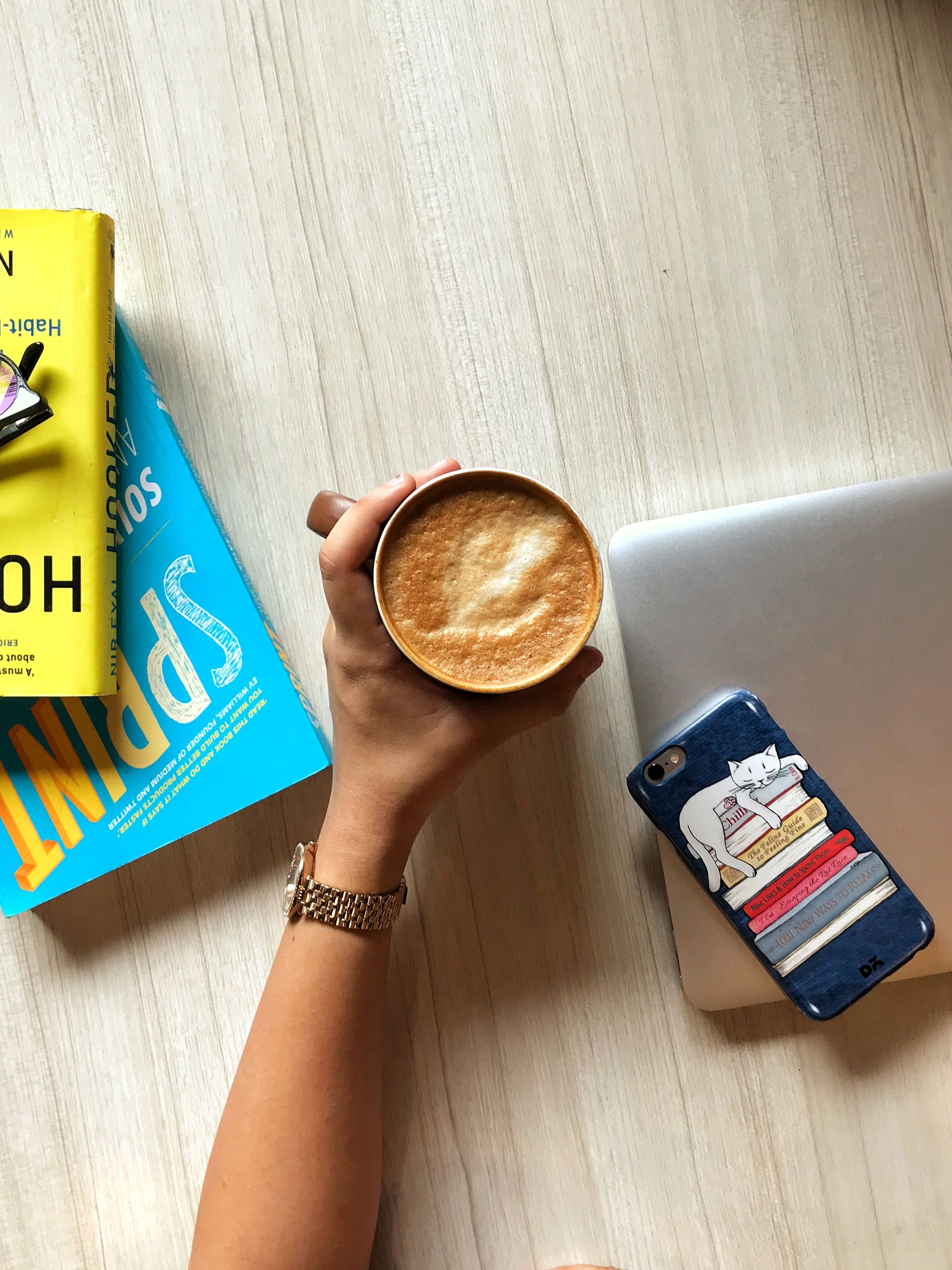Top view of a hand holding a latte on a wooden table with books and a phone nearby.