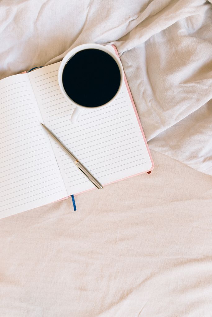 Top view of a lined notebook with a pen and coffee on a bed, evoking cozy morning vibes.