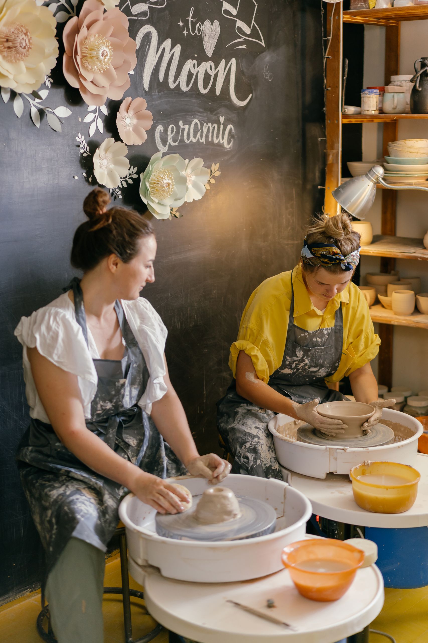 Two women create pottery on wheels in a bright, artistic workshop.