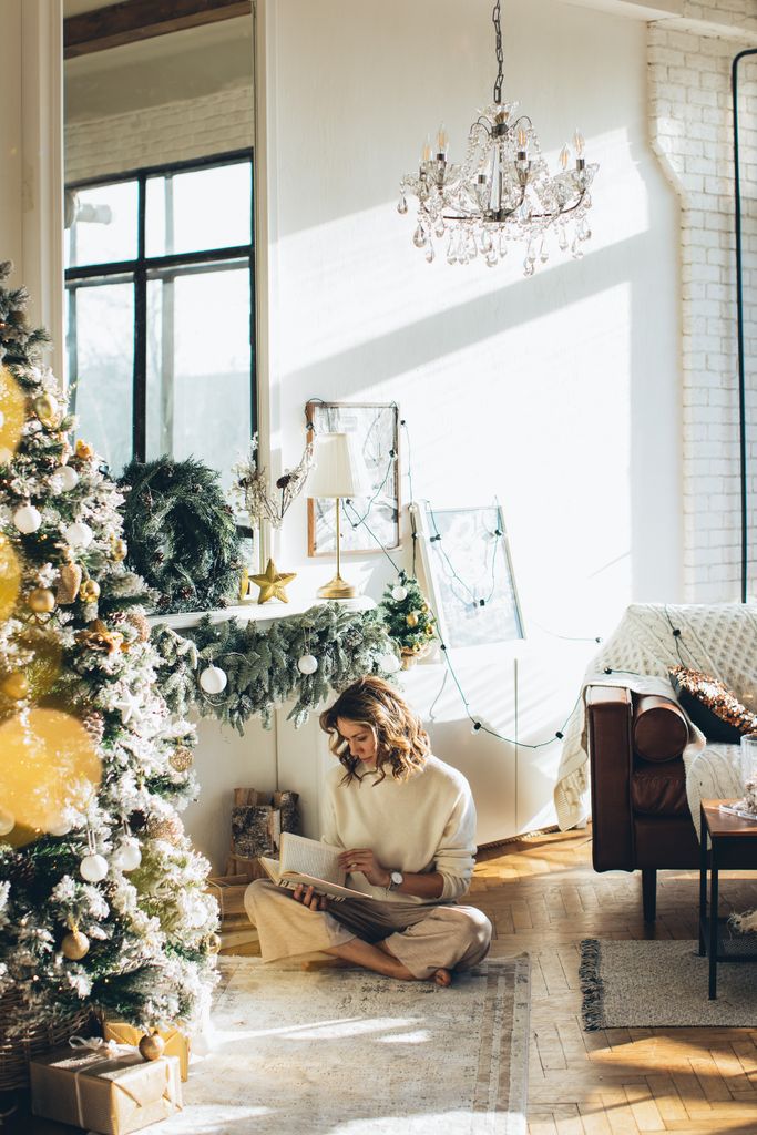 Woman enjoying holiday reading by a Christmas tree in a sunlit, cozy living room.
