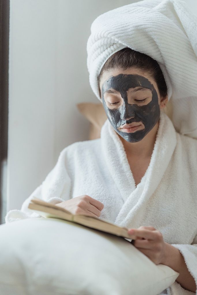 Woman in a bathrobe reading while enjoying a skincare routine with a face mask.