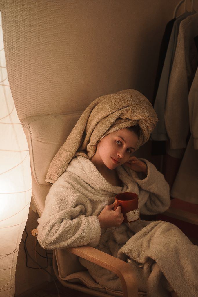 Woman in a bathrobe with a towel on head holding a cup, sitting comfortably indoors.