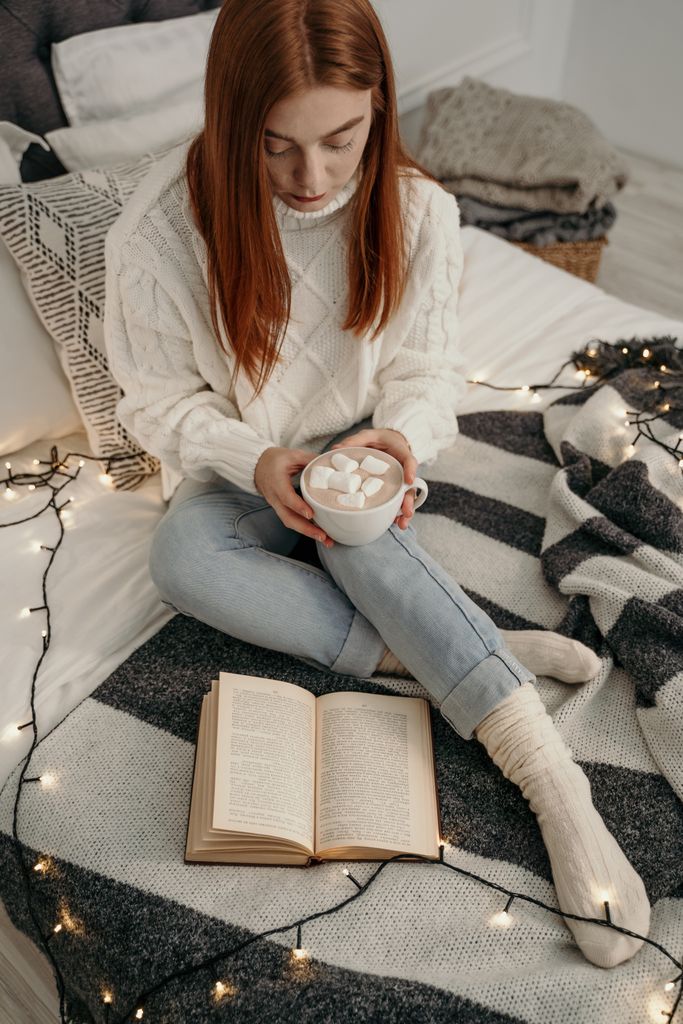 Woman in a cozy knit sweater enjoys hot cocoa while reading on a bed.