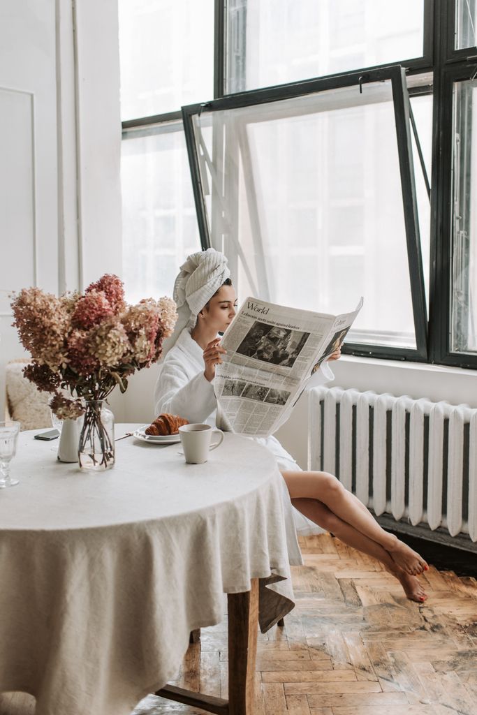 Woman in bathrobe reading newspaper by window, enjoying breakfast with coffee and croissant.millionaire morning routine