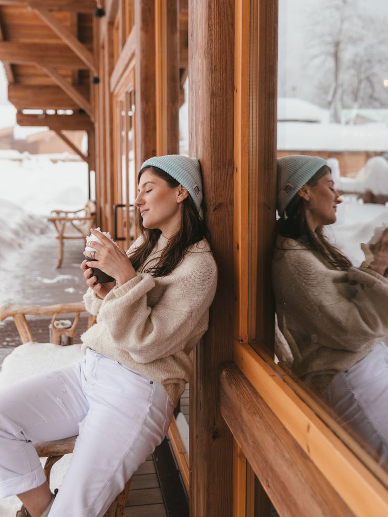 Woman in cozy apparel enjoying a coffee outside a snowy cabin, reflecting tranquility.