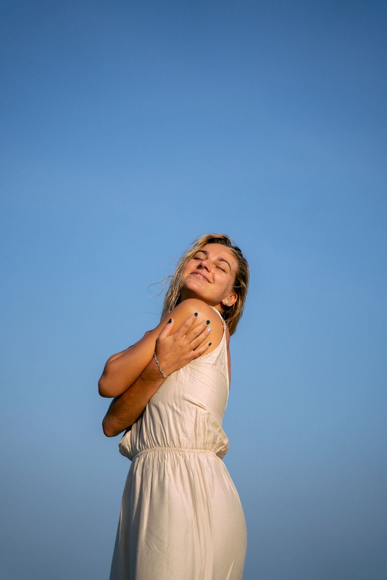 30 day gratitude challenge Woman in white dress embracing sunlight with closed eyes against blue sky.