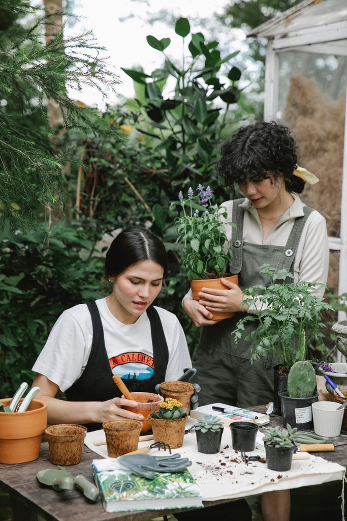 Woman standing with blooming plant near assistant sitting at table and planting seedlings in pots on garden