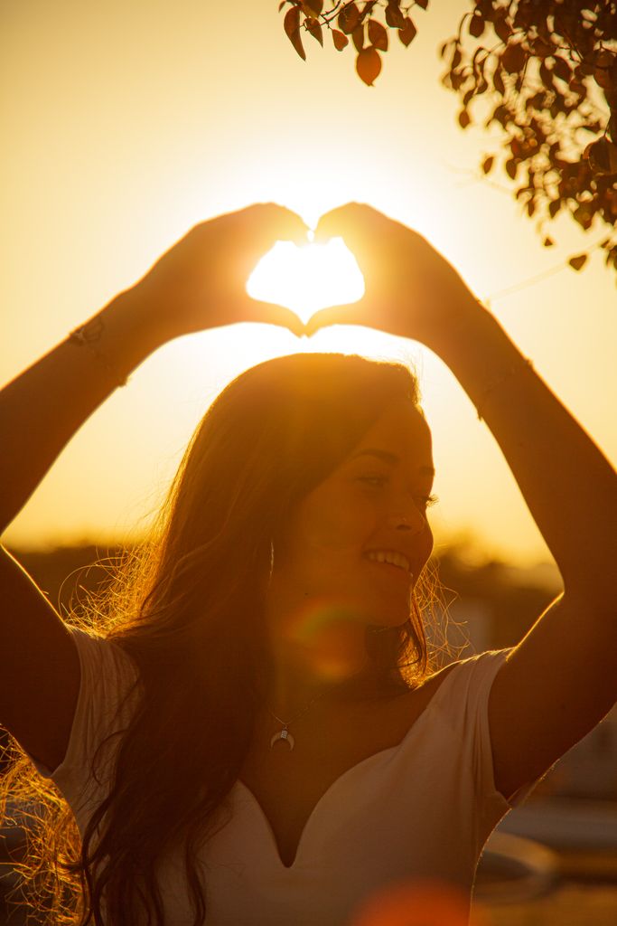 Young cheerful female making heart with hands and smiling while looking away at sundown
