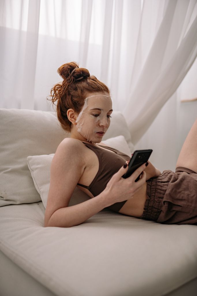Young woman enjoying a self-care skincare routine with a face mask while using her smartphone on a cozy couch.