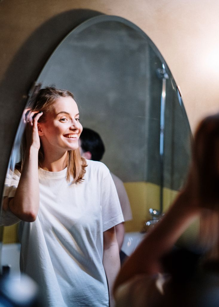 Young woman enjoying her morning routine, smiling at her reflection in a cozy bathroom mirror.